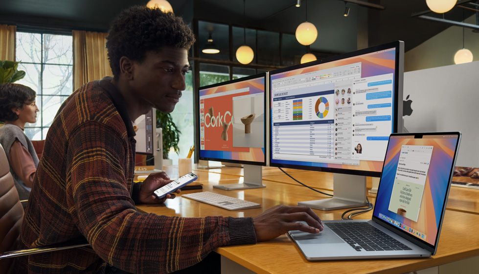 一个在办公环境中的人物，他正在使用一台打开的 MacBook Air，这台电脑连着两台外接显示器和妙控键盘，人物的另一只手里拿着一部 iPhone