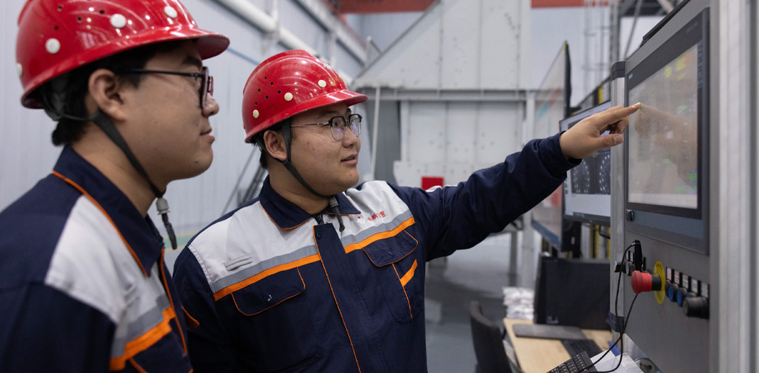 两名戴着安全帽、穿着制服的员工看着数字屏幕，屏幕下方有控制手柄