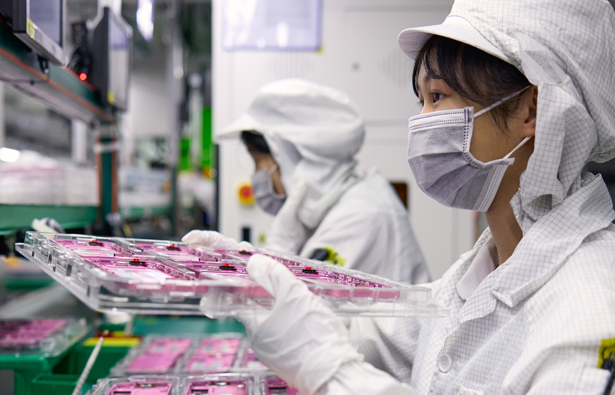 两名穿着全罩式工作服的员工在组装厂处理 iPhone 部件